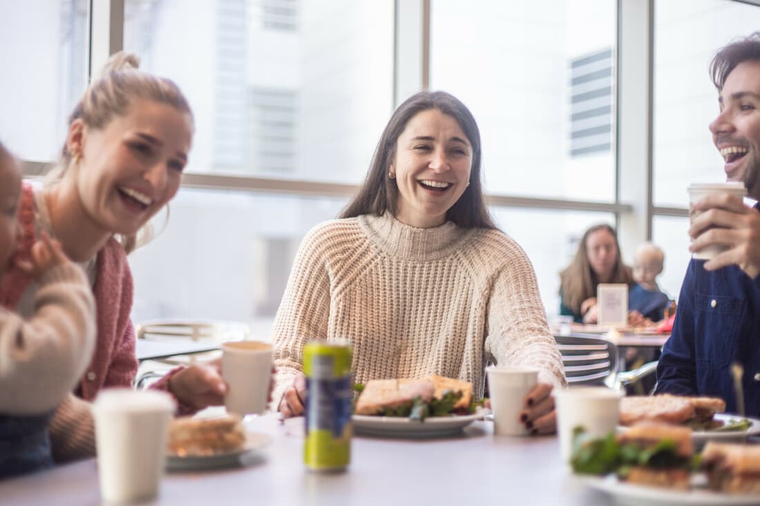 Three adults and a toddler are sitting around a cafe table in Friedman Court, enjoying lunch from the Café and each other's company.